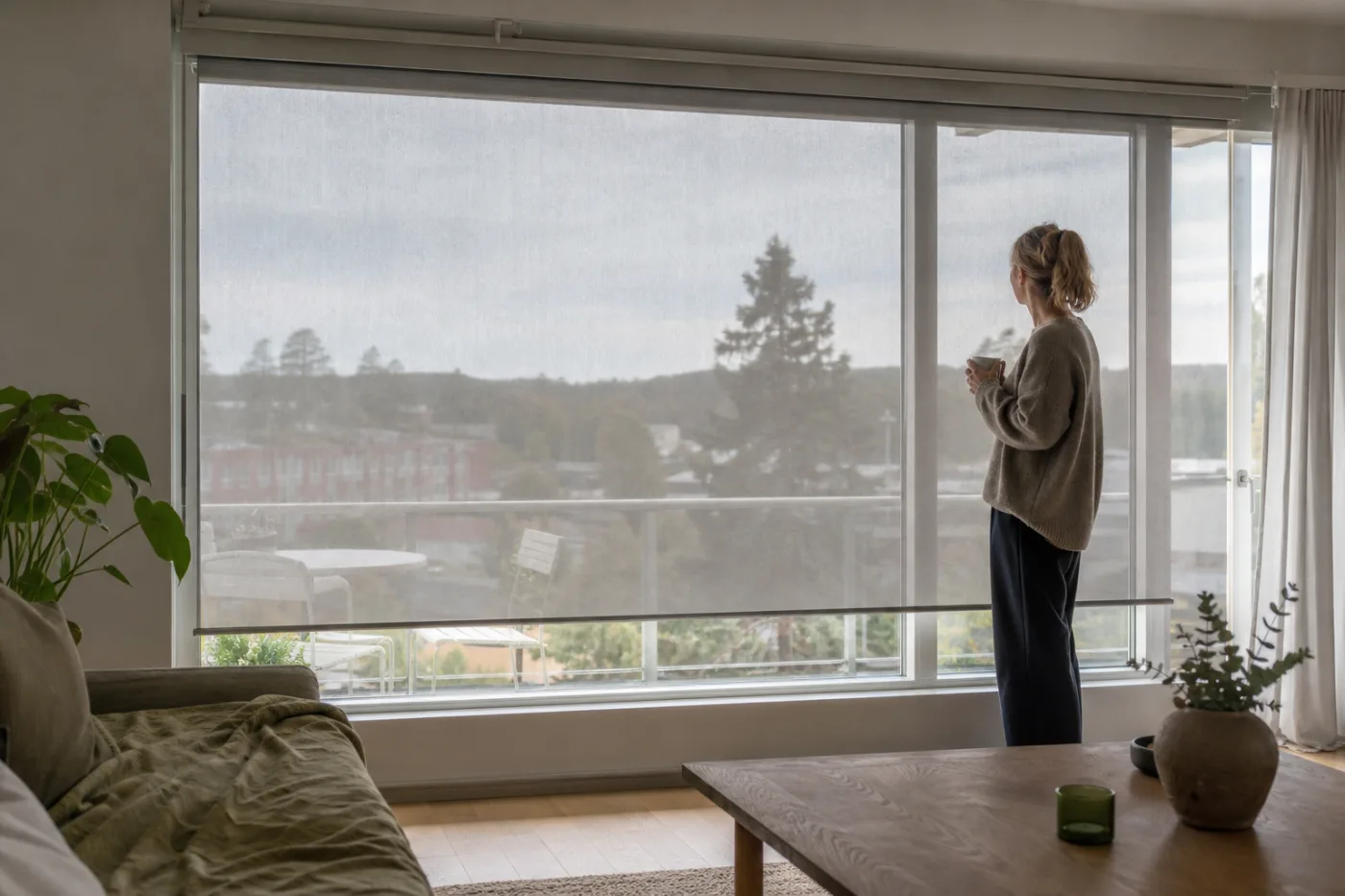 Vardagsrum med stora f&ouml;nster mot balkong d&auml;r en ljusgr&aring; vertikal screenmarkis d&auml;mpar solljuset